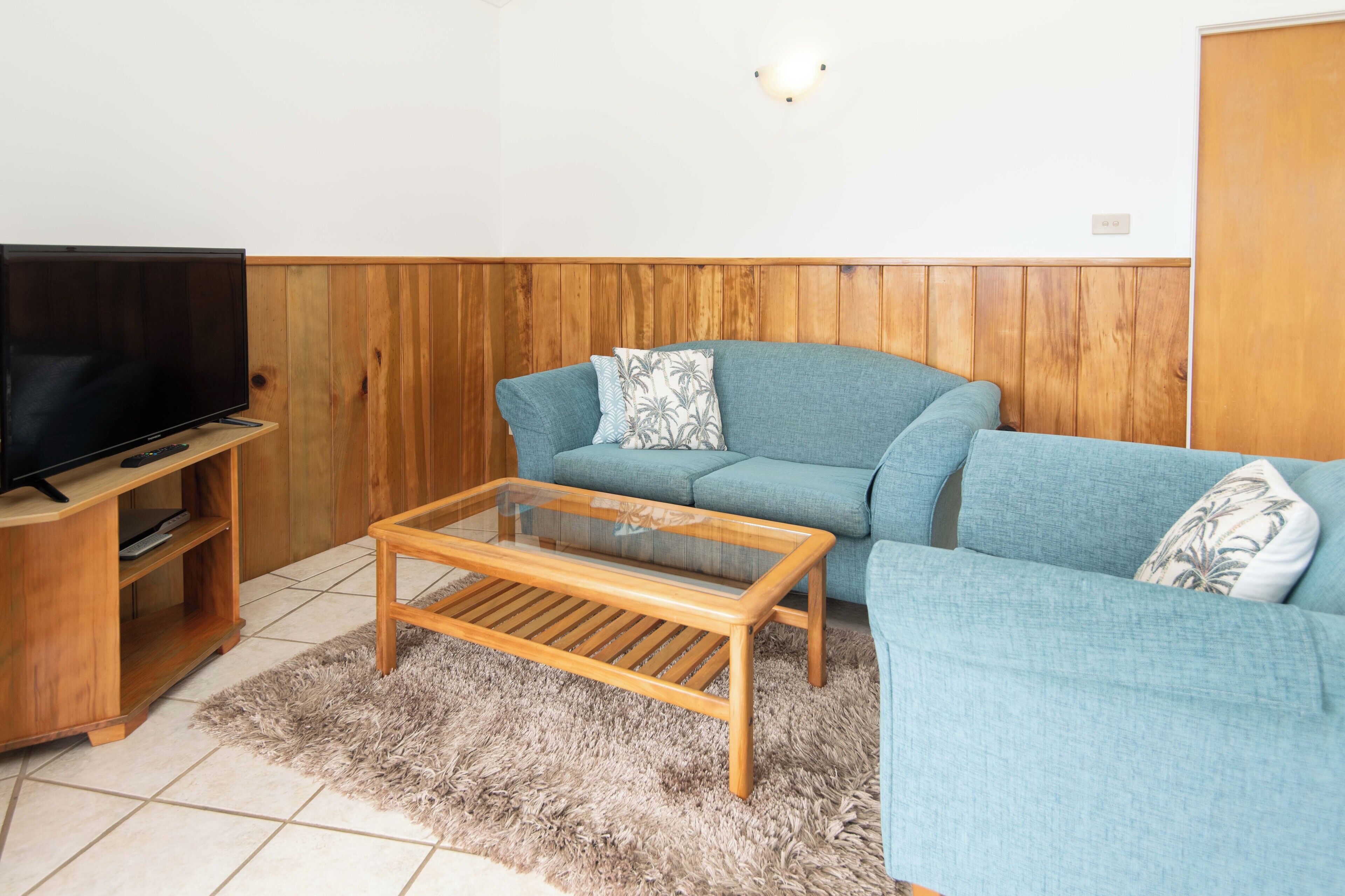 A living room with a television on a wooden stand, a glass-top coffee table, a blue sofa with patterned cushions, and a wooden wall panel.