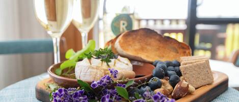 A wooden board with cheese, bread, and flowers, accompanied by two glasses of white wine.