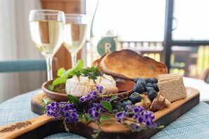 A wooden board with cheese, bread, and flowers, accompanied by two glasses of white wine.