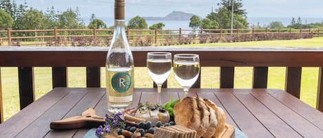 A wooden table with a bottle of rosé wine, two glasses, a cheese board with bread, and a view of the ocean and trees.