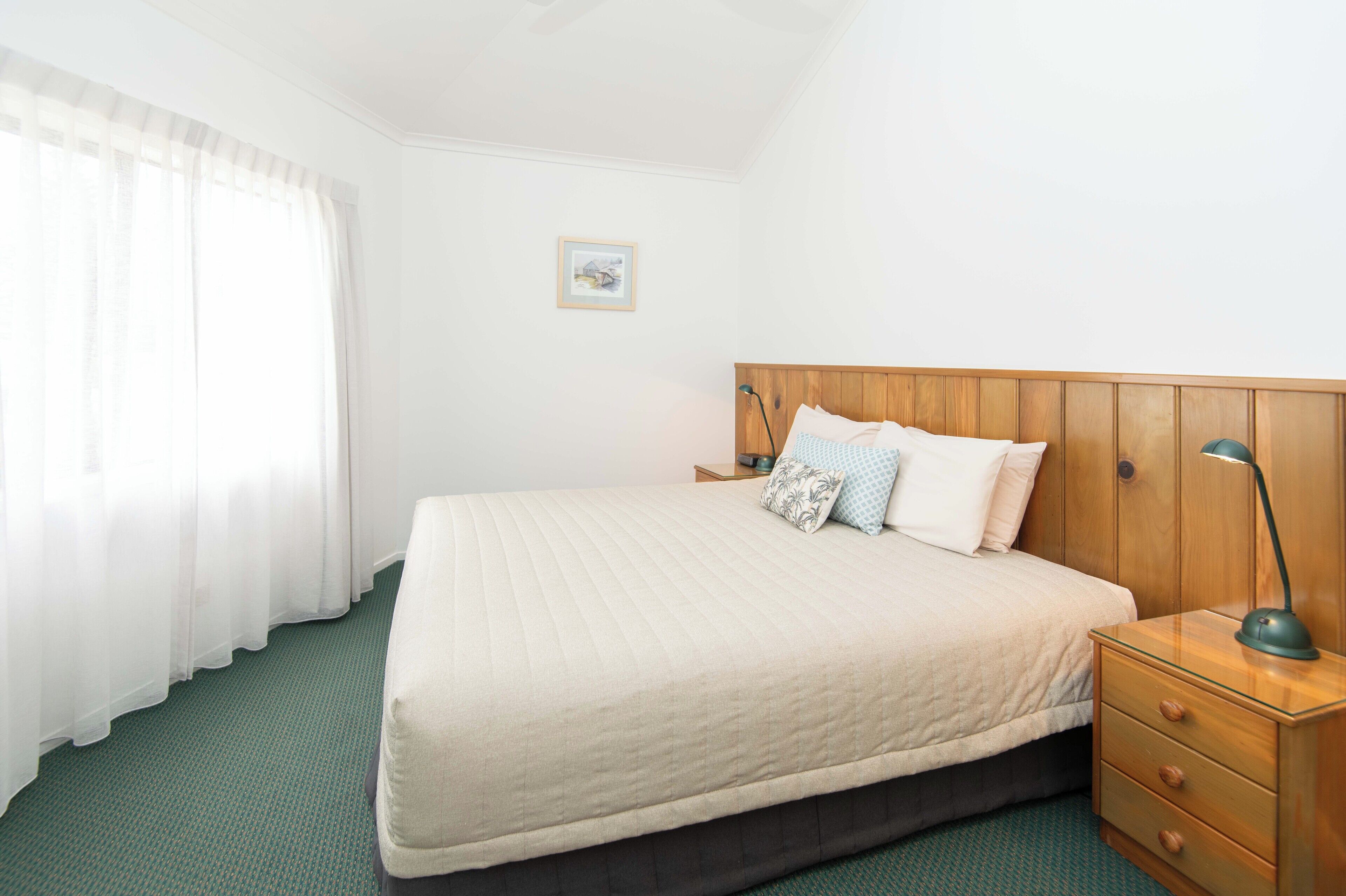 Una cama bien hecha con cabecera de madera, una mesita de noche con lámpara y un suelo cubierto de moqueta verde.
