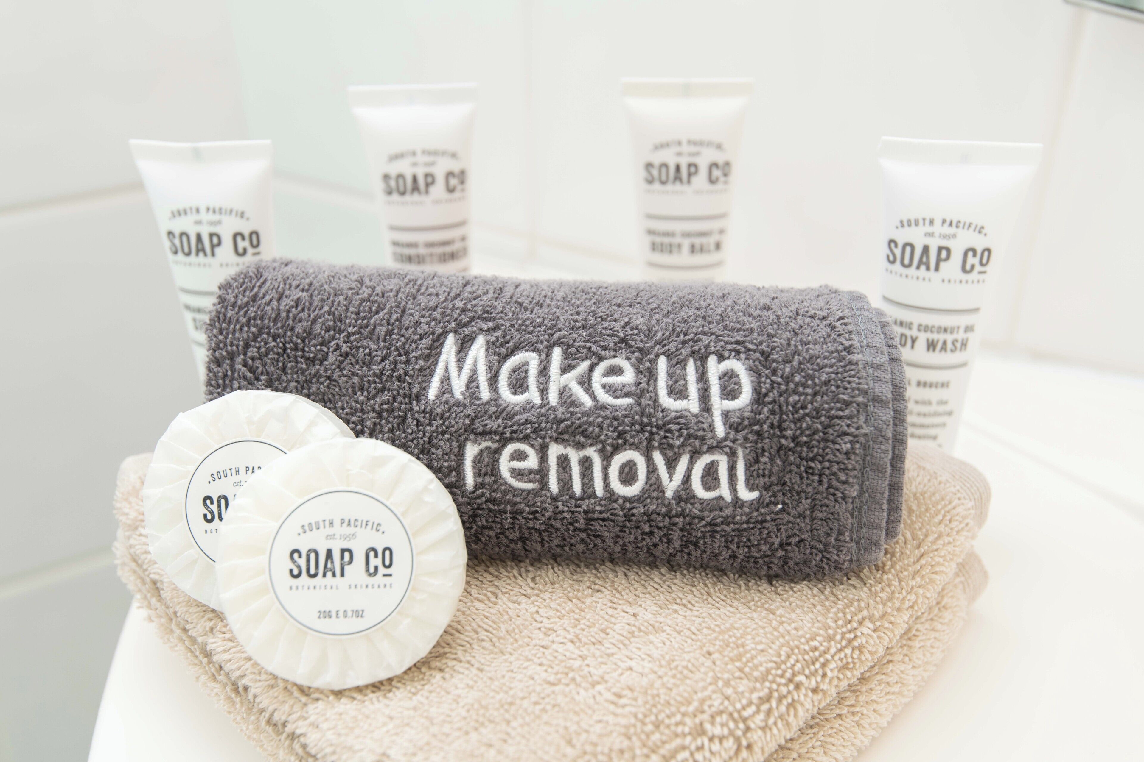 Une salle de bain équipée d’une serviette grise sur laquelle est inscrit « pour enlever le maquillage » et de deux savons ronds présentés dans des contenants portant la marque « SOAP CO ».