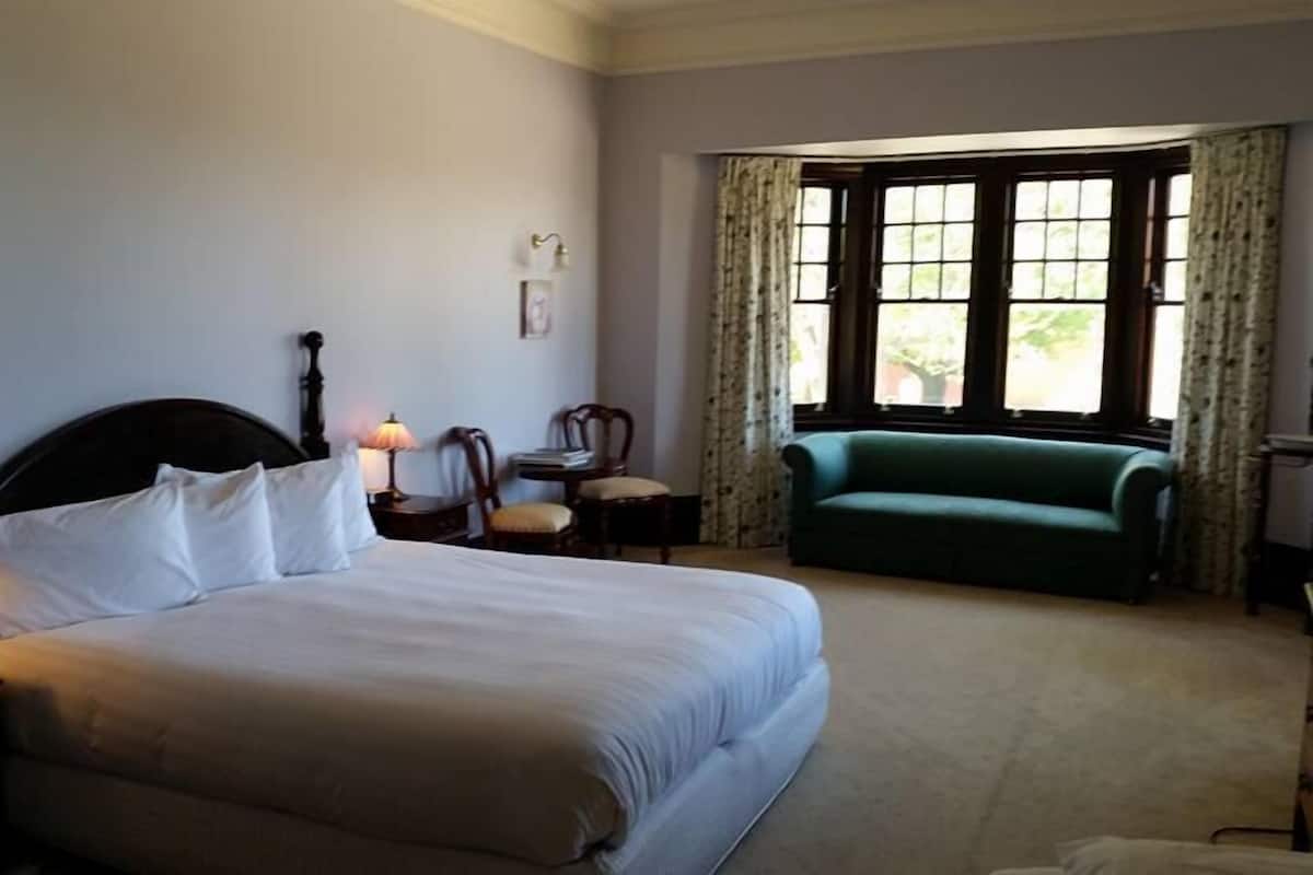 A hotel room with a large bed, a green armchair, a small table, and a window with curtains.