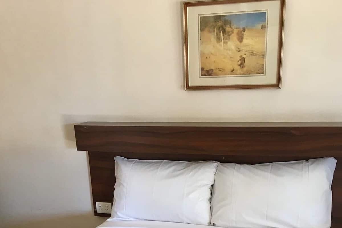 A neatly made bed with a beige bedspread and pillows, a framed painting on the wall, and a power outlet on the headboard.
