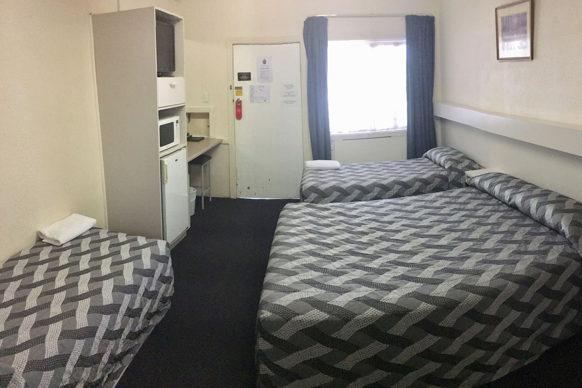 A hotel room with two beds, a kitchenette, a door with a notice, and a window with curtains.