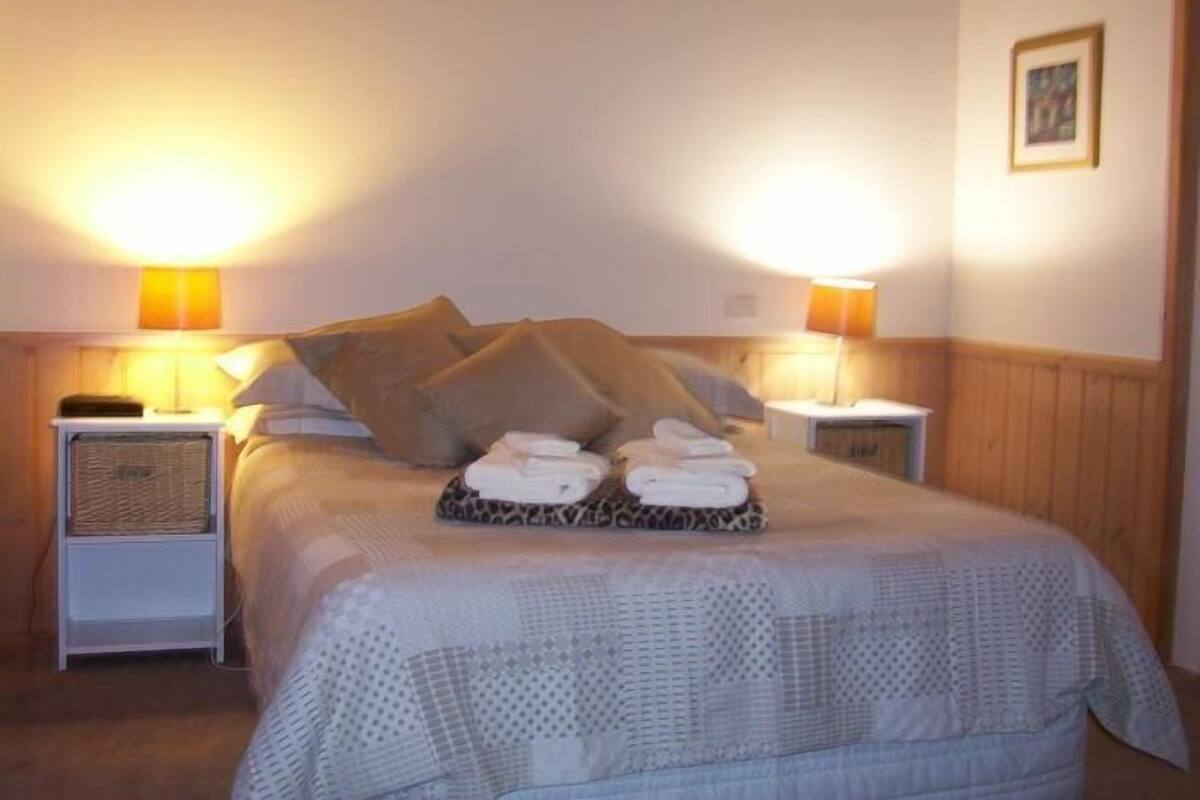 Una cama bien hecha con una colcha estampada, dos almohadas y una pila de toallas dobladas sobre una mesita de noche.