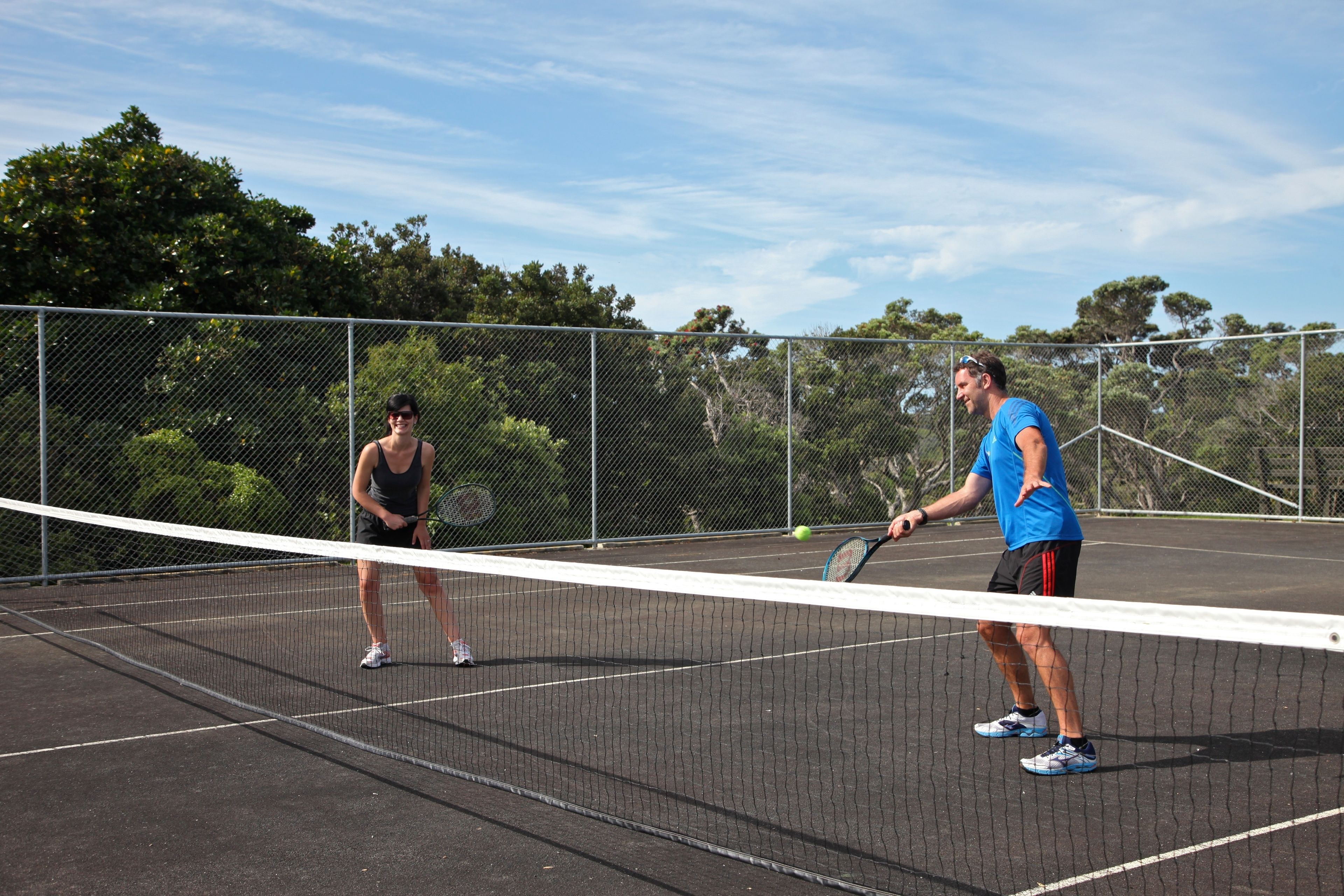 A tennis court with a net in the middle, surrounded by a chain-link fence and trees in the background.