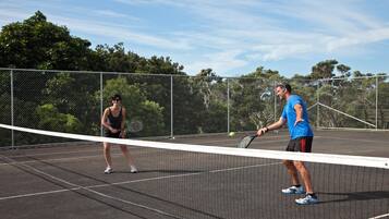 A tennis court with a net in the middle, surrounded by a chain-link fence and trees in the background.