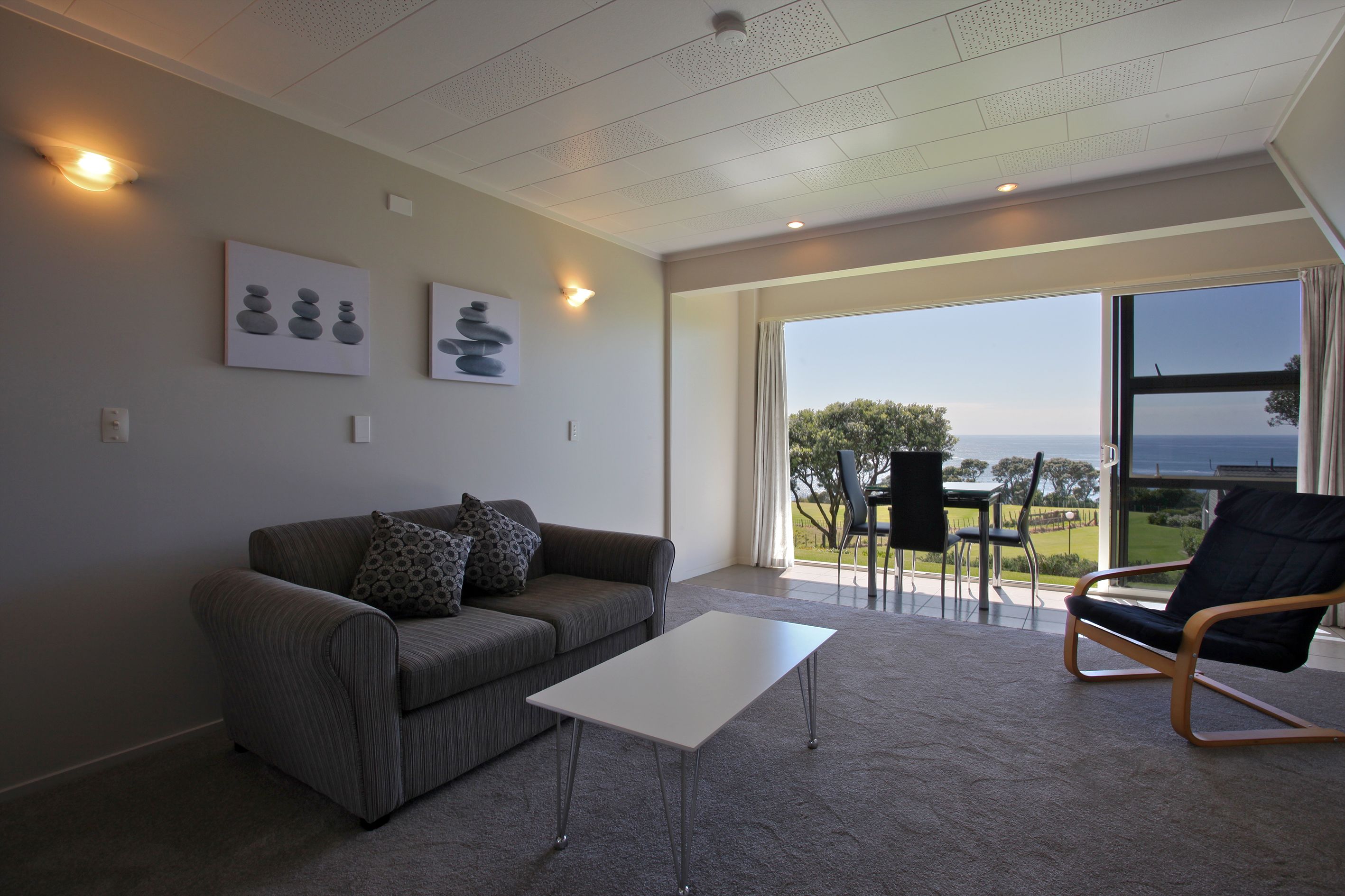 A modern living room with a sofa, a coffee table, and a chair. There are wall-mounted artworks and a large window offering a view of the outdoors.