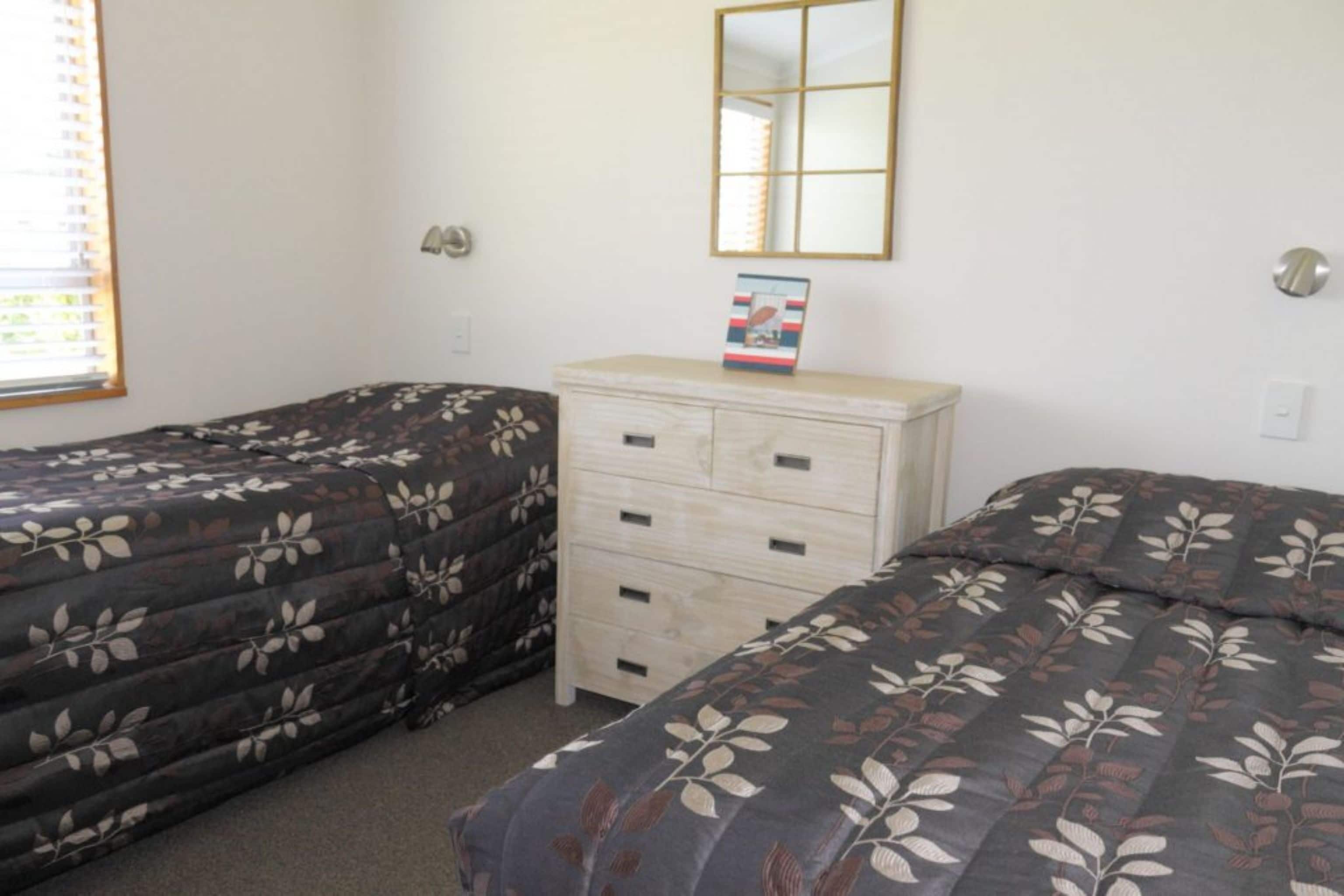 A room with two single beds, a dresser, a mirror, and a window with blinds.