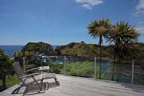 Una terraza de madera con dos sillas y una mesita pequeña, con vistas a un paisaje costero que incluye el mar y follaje.
