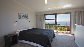 A bedroom with a large bed, a nightstand, a painting, a view of the sea through a large window, and a balcony with chairs.