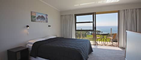 A bedroom with a large bed, a nightstand, a painting, a view of the sea through a large window, and a balcony with chairs.