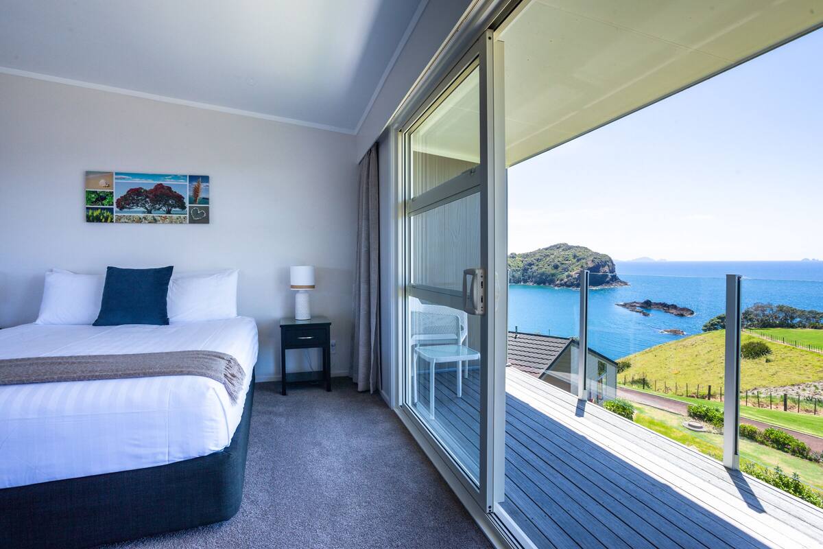A bedroom with a bed, a nightstand, a lamp, and a large window with a view of the sea and a grassy area.