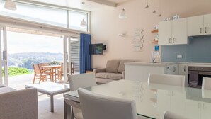 A modern living room with a glass dining table, white sofas, and a kitchen area with white cabinets and a blue countertop.