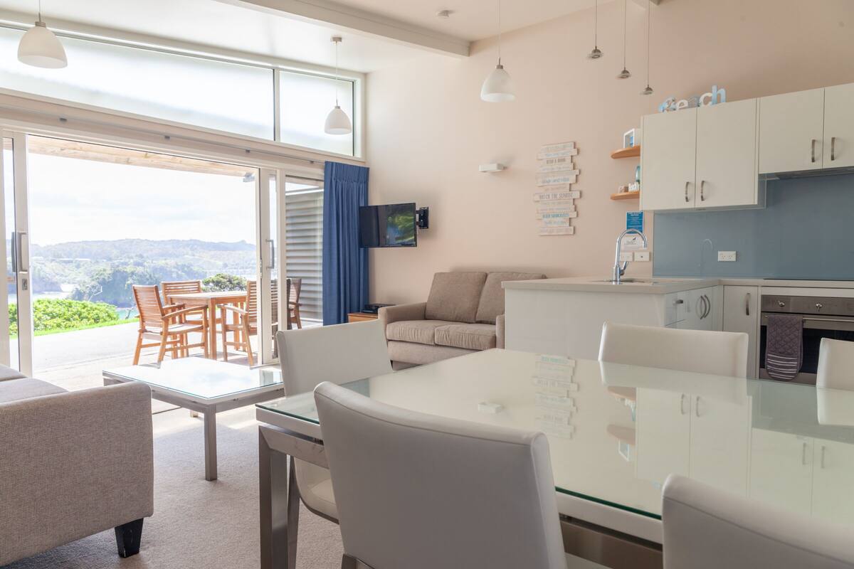 A modern living room with a glass dining table, white sofas, and a kitchen area with white cabinets and a blue countertop.