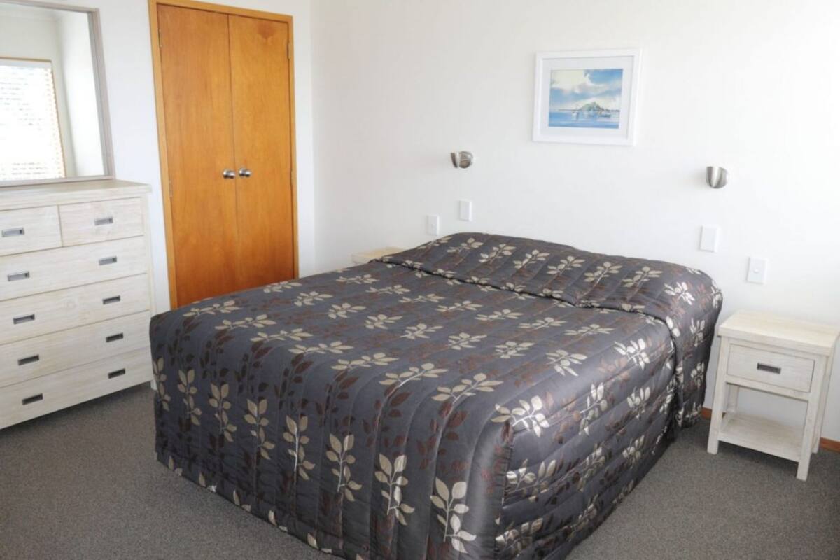 A bedroom with a bed, a dresser, a nightstand, a door, and a framed picture on the wall.