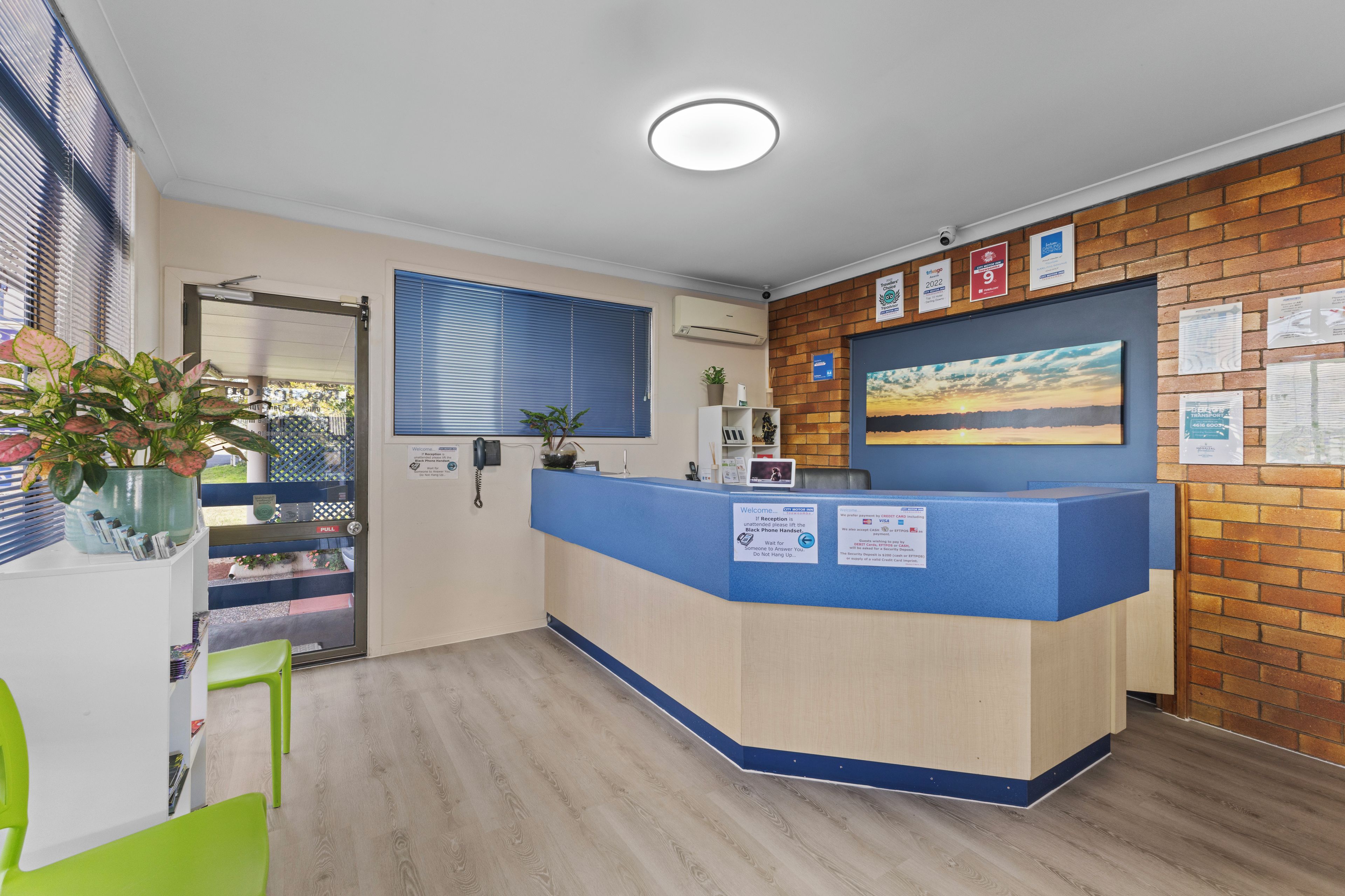 Una recepción con mostrador azul, una planta en maceta, una ventana con persianas y una pared con carteles informativos.