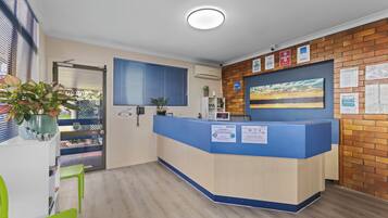 Una recepción con mostrador azul, una planta en maceta, una ventana con persianas y una pared con carteles informativos.