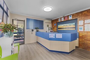 A reception desk with a blue counter, a potted plant, a window with blinds, and a wall with informational posters. - City Motor Inn Toowoomba (South Toowoomba)