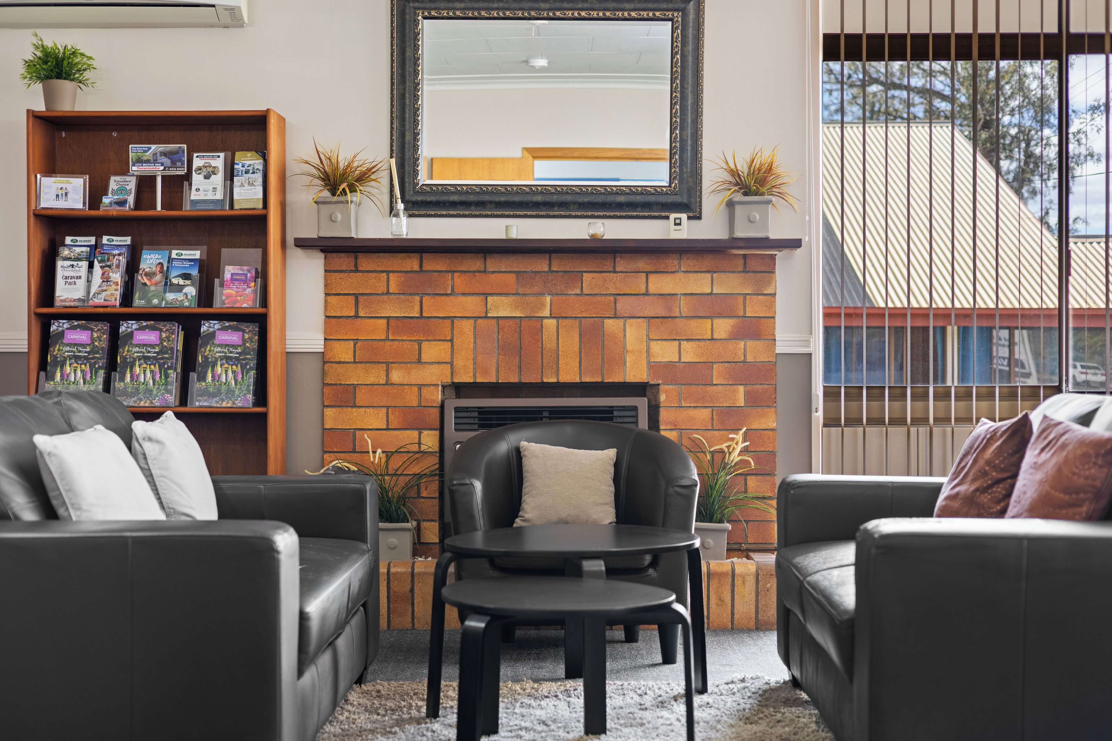 A living room with a brick fireplace, black leather sofas, a small table, and a bookshelf with travel brochures.