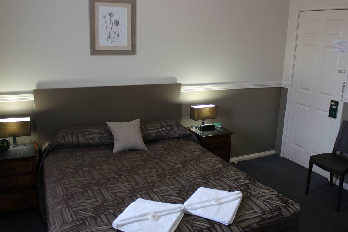 A neatly made bed with a patterned bedspread, two pillows, a bedside lamp, a nightstand, and a framed picture on the wall.