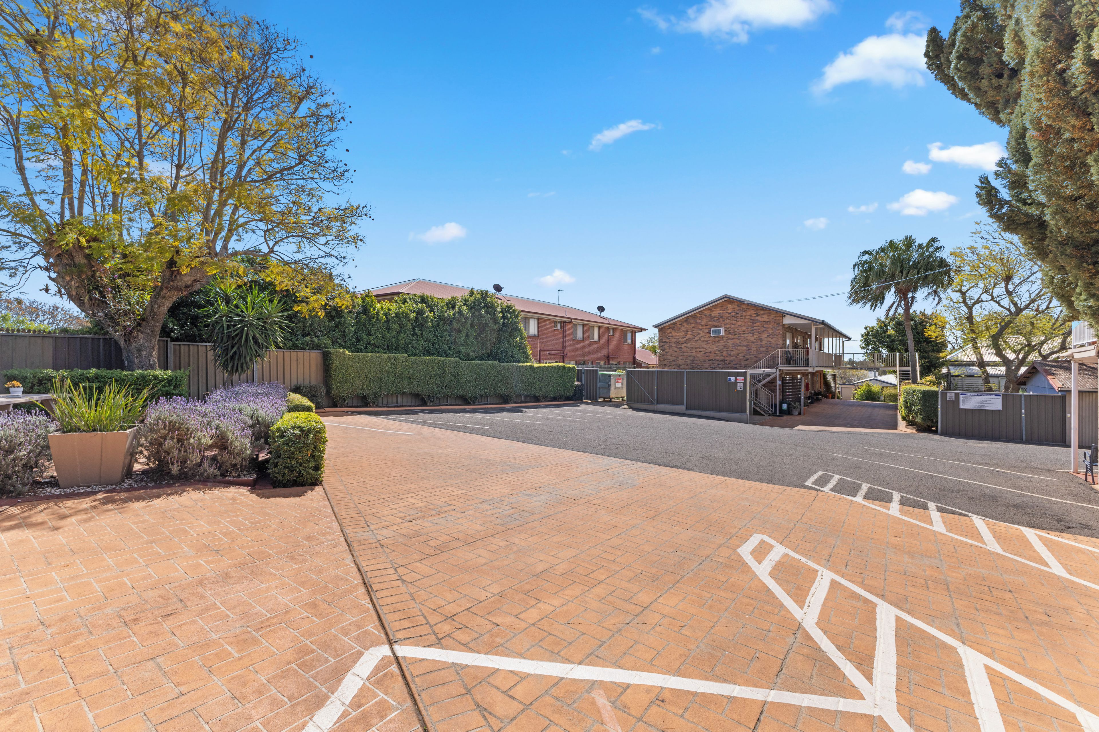 A paved driveway leading to a gated entrance with a brick building and a large tree.