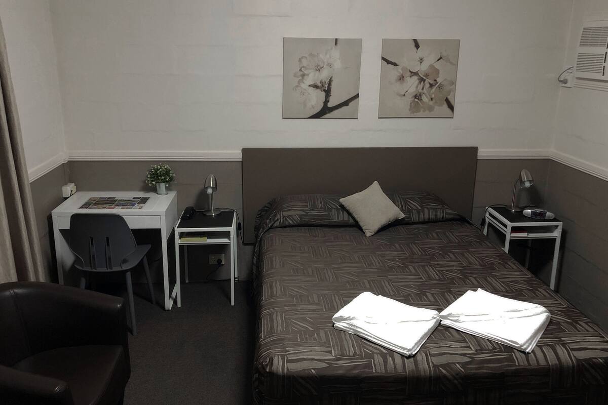 A hotel room with a single bed, a desk, a chair, and two wall-mounted paintings.
