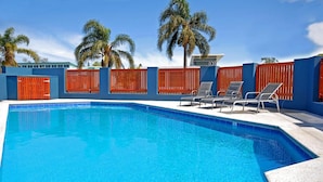 A swimming pool with blue water, surrounded by a blue wall and red fences. There are two lounge chairs and a palm tree in the background. - East Port Motor Inn (Port Macquarie)