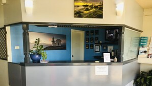 A reception desk with a blue wall, a framed picture of a lighthouse, and a sign indicating the reception hours. - East Port Motor Inn (Port Macquarie)