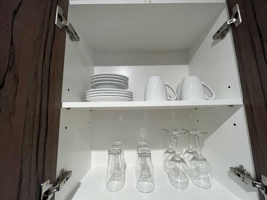 A white shelf unit with two compartments; the upper shelf holds stacked plates and white mugs, while the lower shelf contains clear wine glasses and a single bottle.