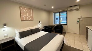 A hotel room with a bed, a desk, a chair, a window with a view, and an air conditioning unit. - Ryley Motor Inn (Wangaratta)