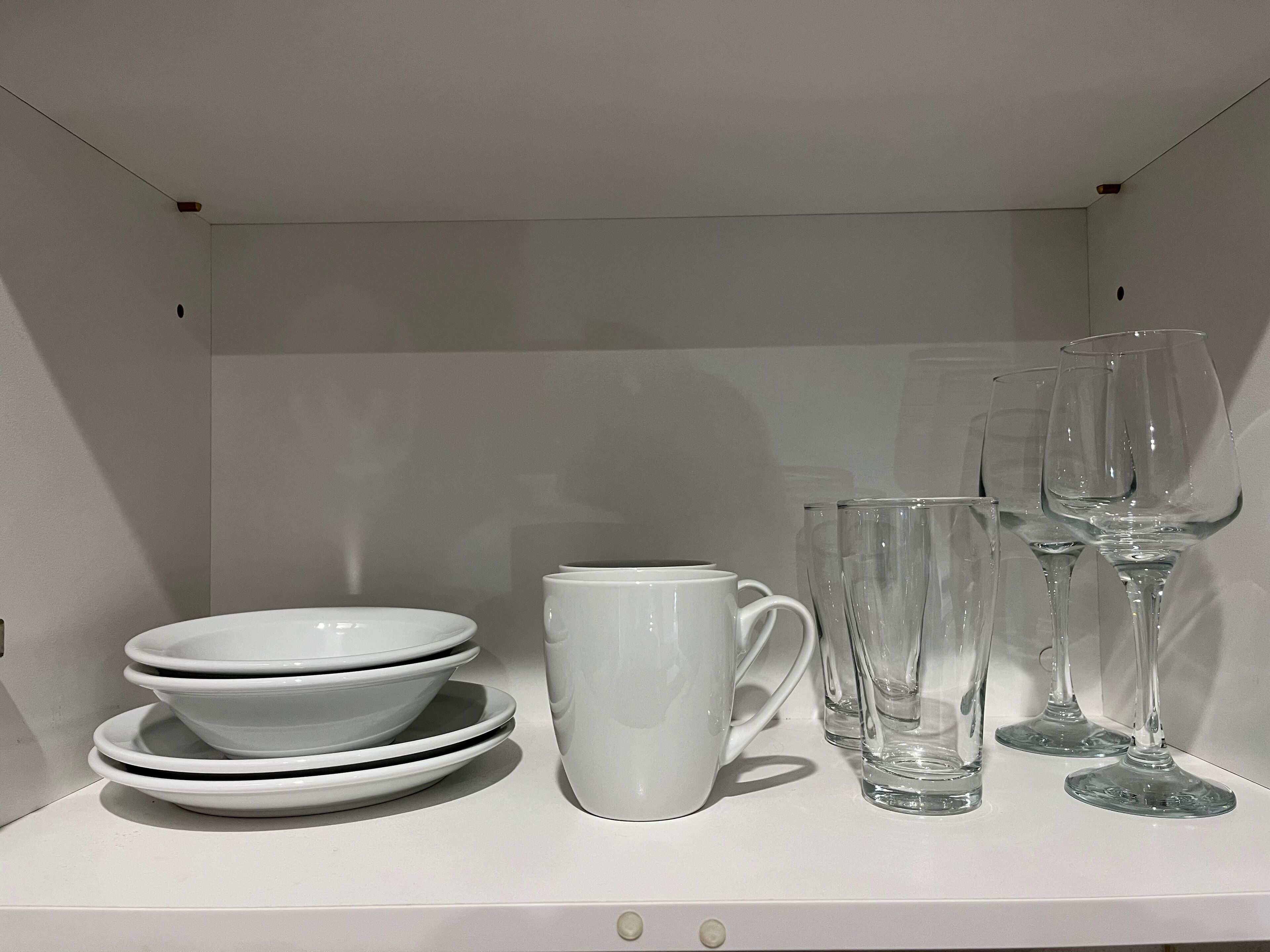 A white shelf with stacked white bowls, a white mug, and clear wine glasses.