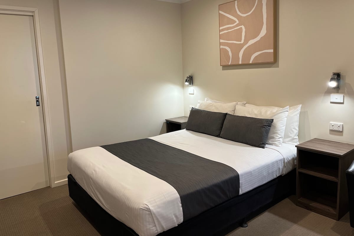 Quarto de hotel com uma cama grande, duas mesas de cabeceira, um criado-mudo, uma luz acoplada à parede e uma porta.