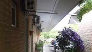 Un couloir extérieur abrité, avec un banc, des plantes en pot et un chemin menant à l’entrée d’un bâtiment.