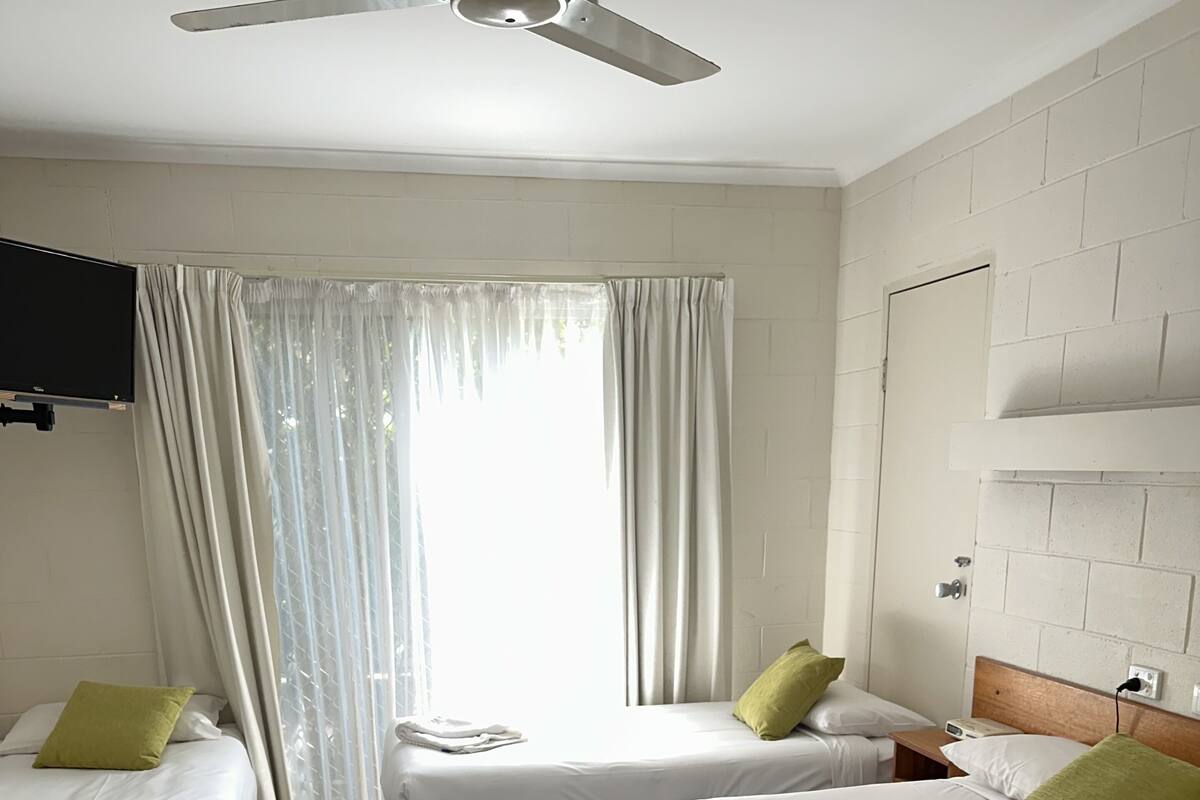 Habitación de hotel con dos camas, ventilador de techo, televisor y ventana con cortinas.