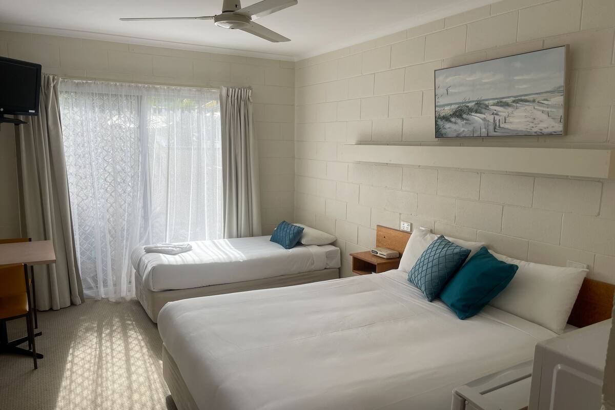 Una habitación de hotel con dos camas, un ventilador de techo, un cuadro en la pared y una ventana con cortinas.
