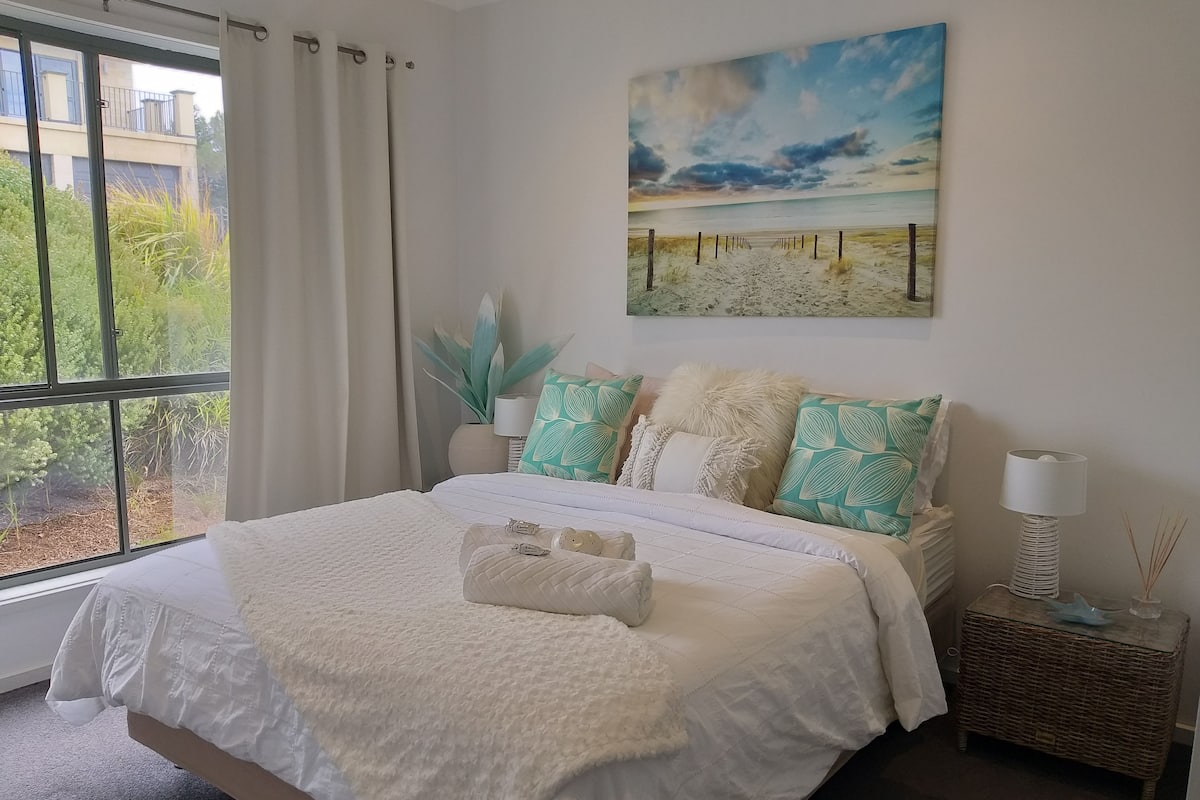 A neatly made bed with a white duvet, a bedside lamp, and a painting of a beach scene.
