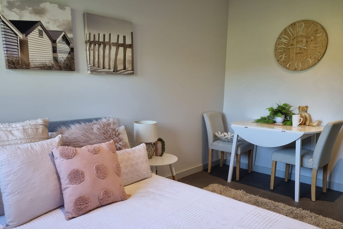 A bedroom with a bed, pillows, a bedside table with a lamp, a chair, a small table with a plant, and a wall clock.