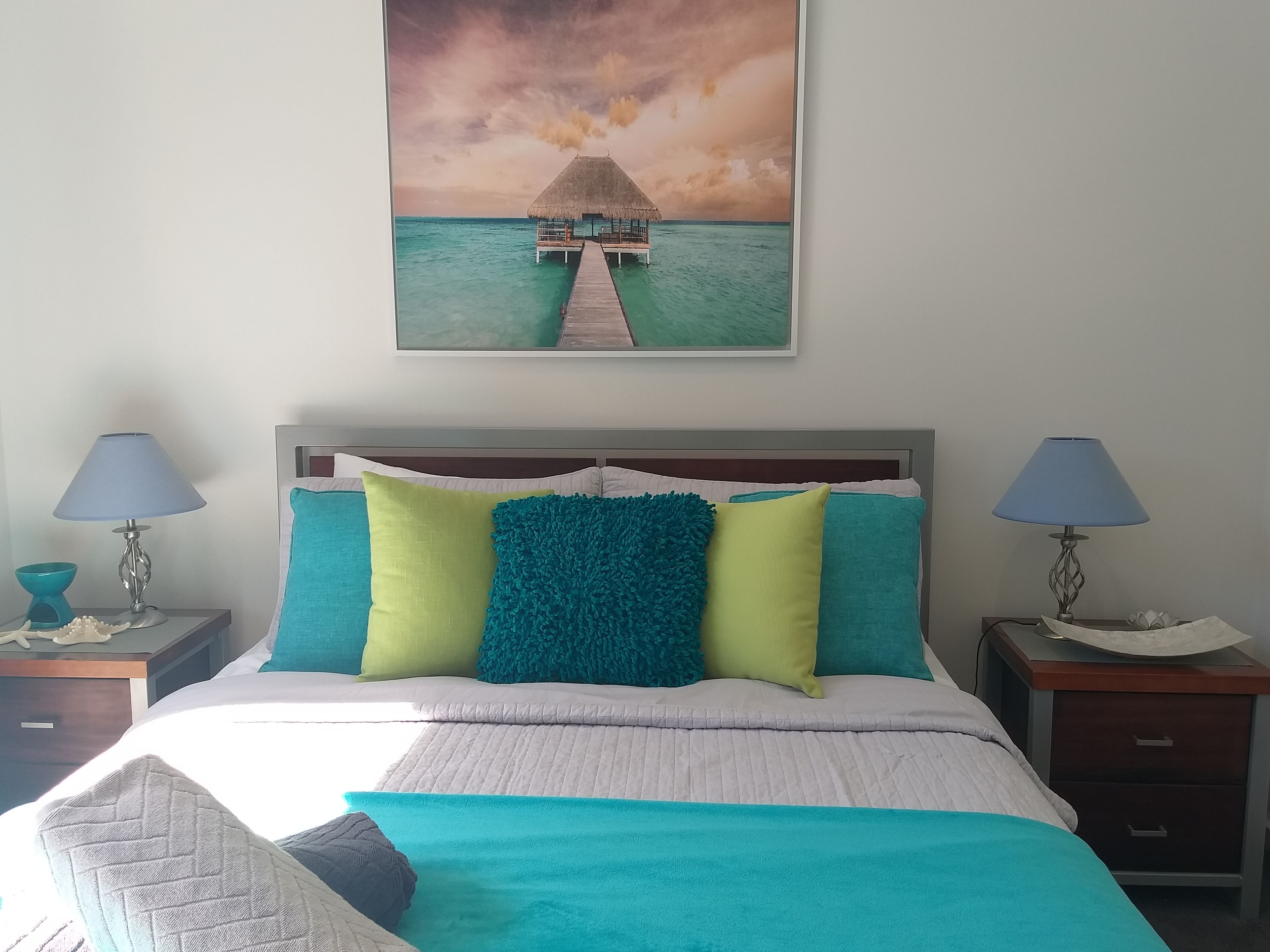 Un dormitorio con cama, dos mesitas de noche, una mesilla auxiliar y un cuadro colgado en la pared que representa una escena tropical.