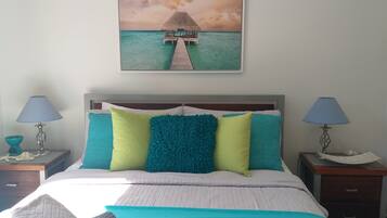 Un dormitorio con cama, dos mesitas de noche, una mesilla auxiliar y un cuadro colgado en la pared que representa una escena tropical.