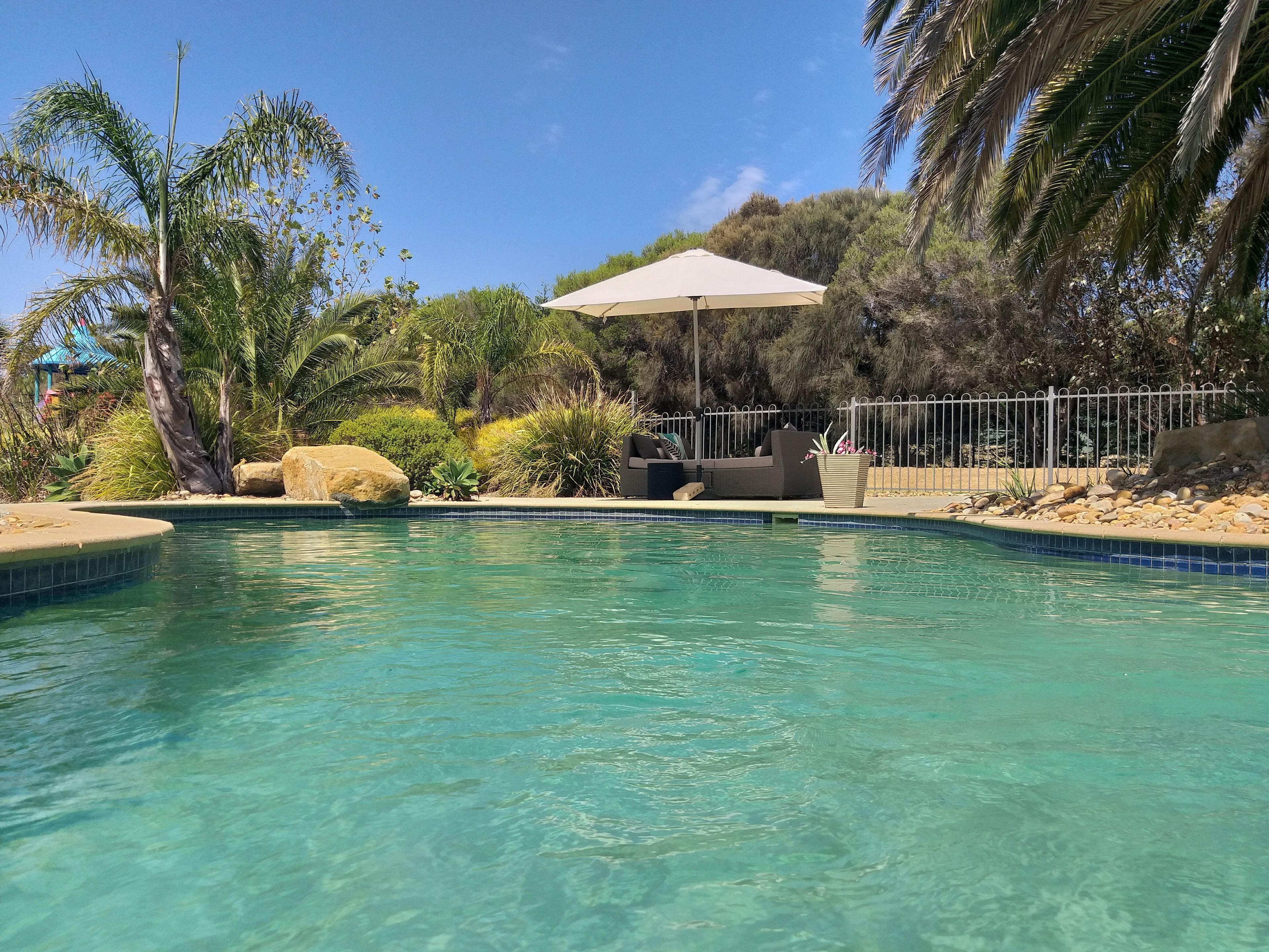 Piscina cristalina com área de estar, cercada por palmeiras e pedras.