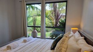 Una cama bien hecha con una colcha blanca y una toalla enrollada, una ventana con vistas a la vegetación, y una mesita de noche con una lámpara.