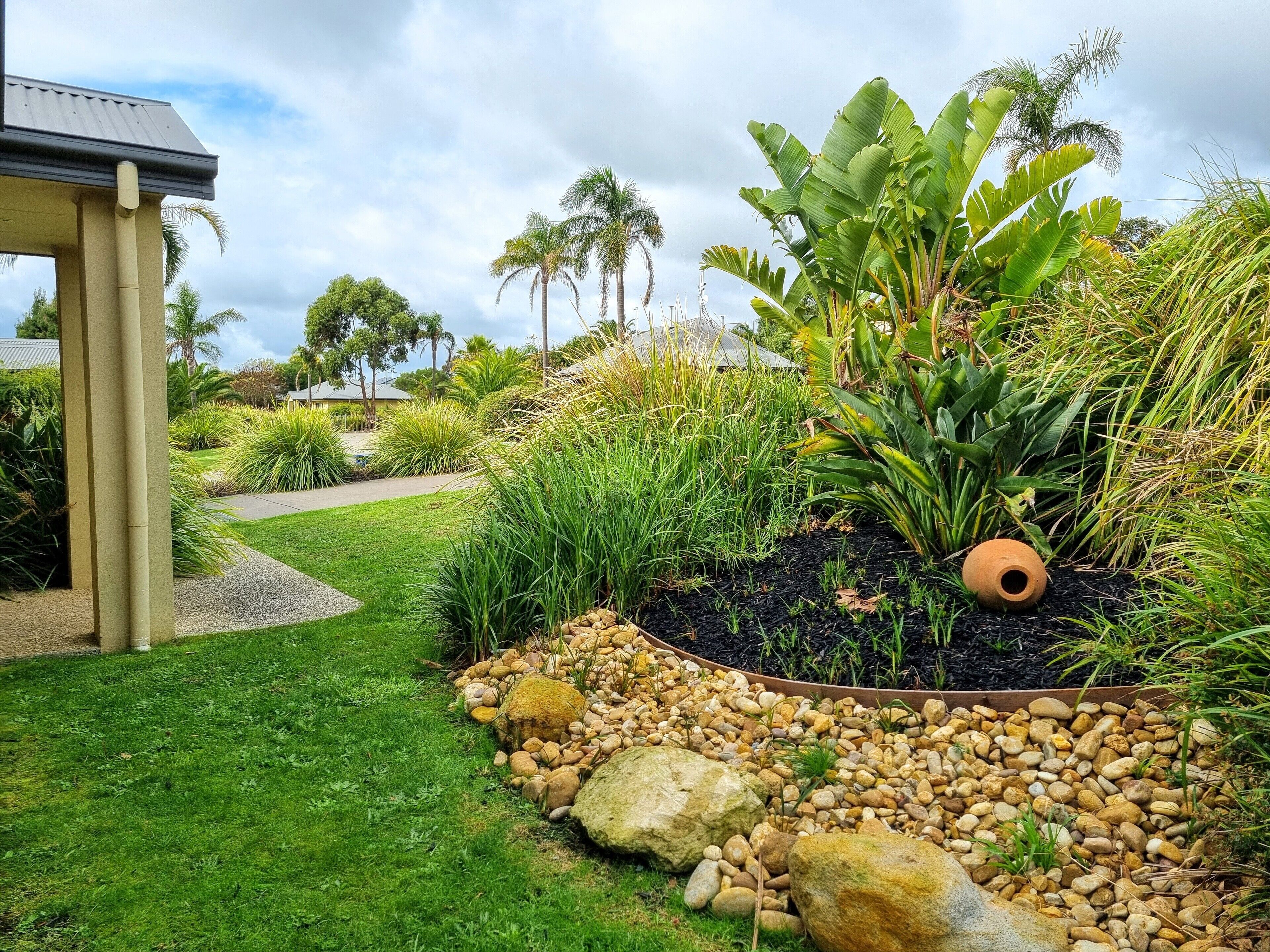 Um jardim com paisagismo, com diversas plantas, incluindo palmeiras e bananeiras, um caminho e uma estrutura com teto.