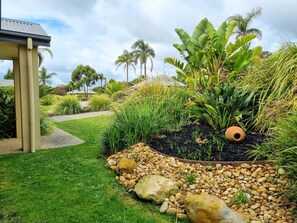 Um jardim com paisagismo, com diversas plantas, incluindo palmeiras e bananeiras, um caminho e uma estrutura com teto.