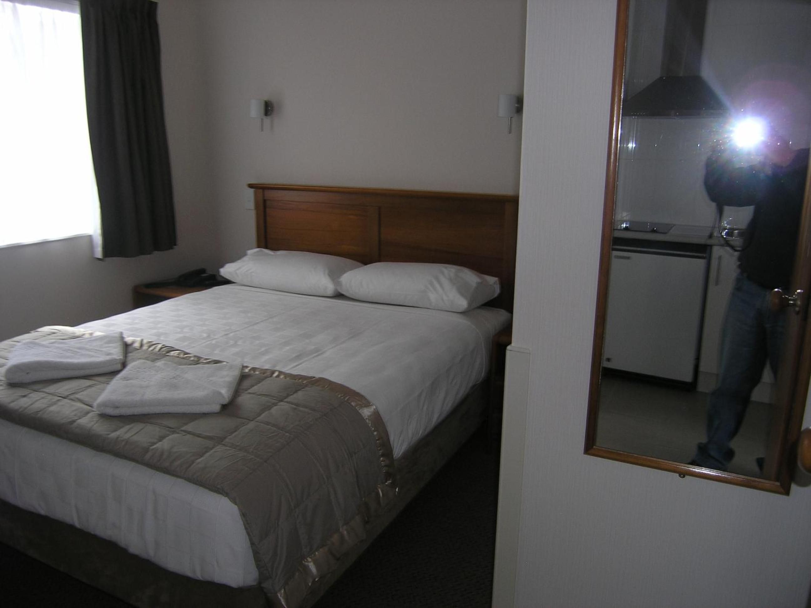 Habitación de hotel con cama, mesita de noche, espejo y kitchenette.