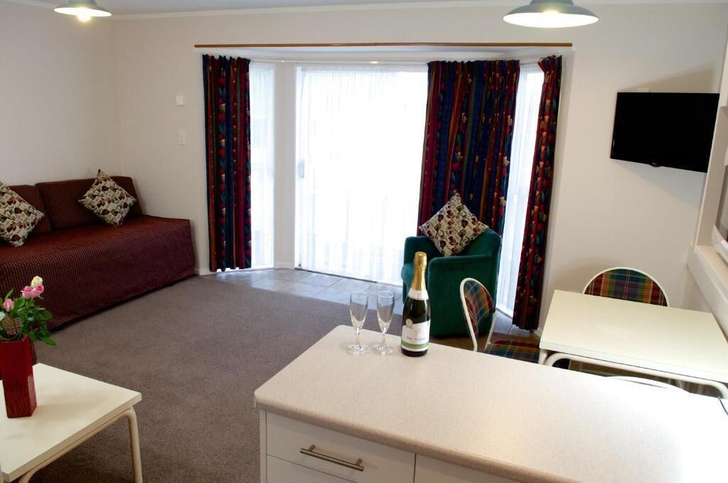 A hotel room with a sofa, a small table with a bottle and glasses, a chair, and a television on the wall.
