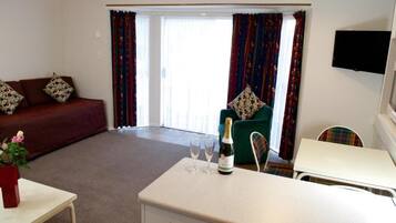 A hotel room with a sofa, a small table with a bottle and glasses, a chair, and a television on the wall.