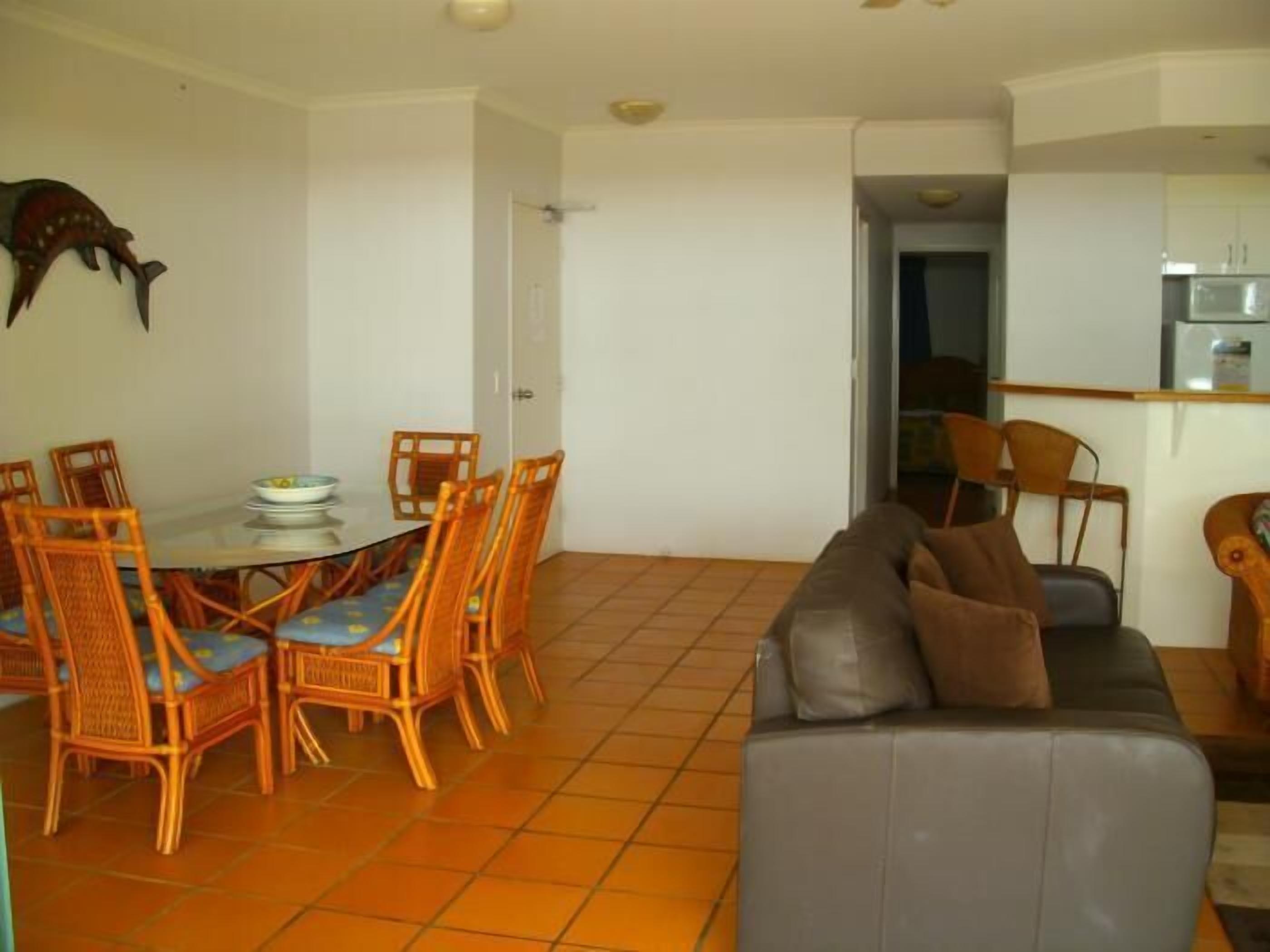 Een eetruimte met een ronde tafel en zes rieten stoelen, een grijze bank met kussens, en een keuken zichtbaar door een open deur.