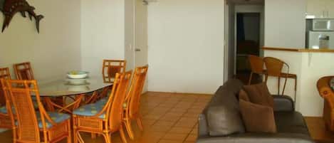 Een eetruimte met een ronde tafel en zes rieten stoelen, een grijze bank met kussens, en een keuken zichtbaar door een open deur.